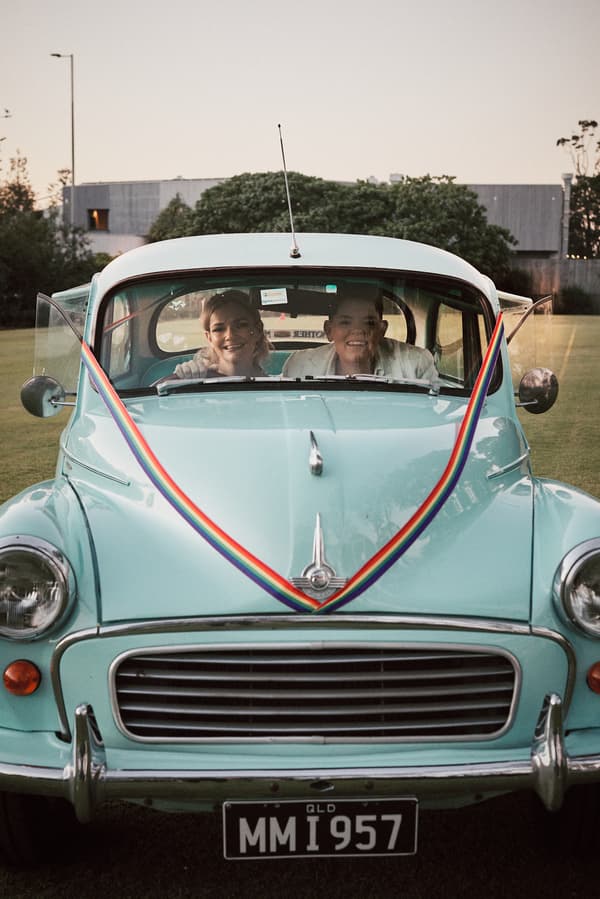 Mandy and Selina sit together inside a vintage light blue car decorated with rainbow ribbons, parked on grass outdoors.