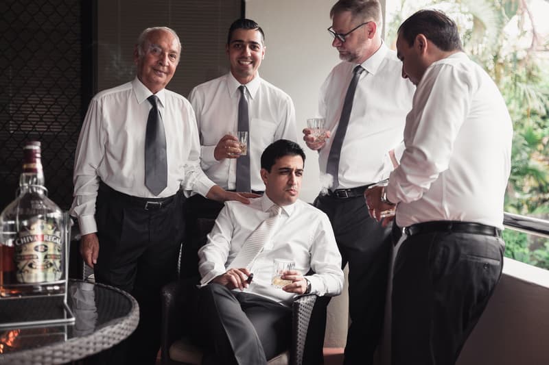 The groom Pasha sits holding a glass and cigar, surrounded by four men in white shirts and ties, standing and holding drinks, near a table with a bottle of Chivas Regal whisky at Hillstone St Lucia — The Quartyard.