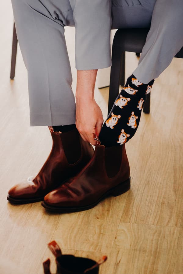 The groom at Sandstone Point Hotel puts on brown leather boots over black socks with a corgi dog pattern while seated on a chair.