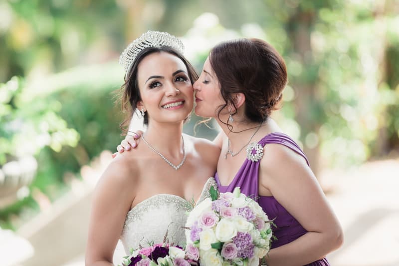 The bride wearing a tiara and strapless wedding gown holds a bouquet while a bridesmaid in a purple dress kisses her on the cheek at Hillstone St Lucia — The Quartyard.