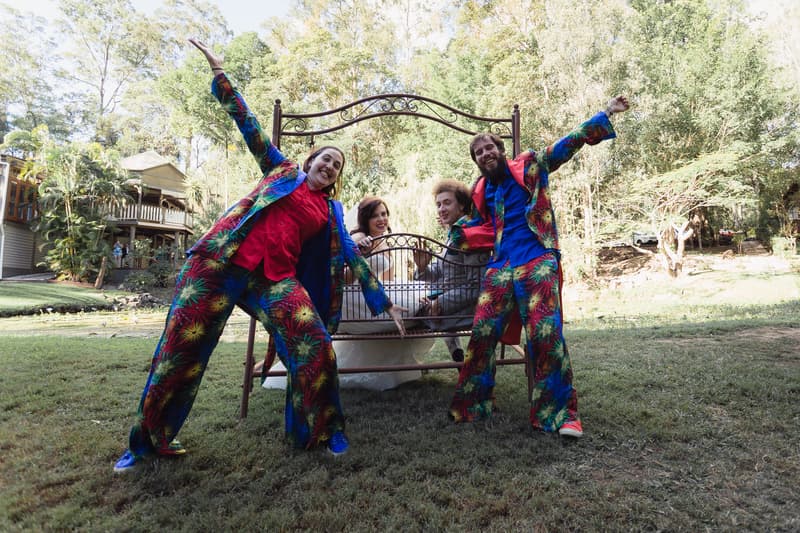 The bride Holly and groom Paul sit on a decorative metal bench outdoors at Kwila Lodge, with two groomsmen in colorful patterned suits posing energetically in front of them.