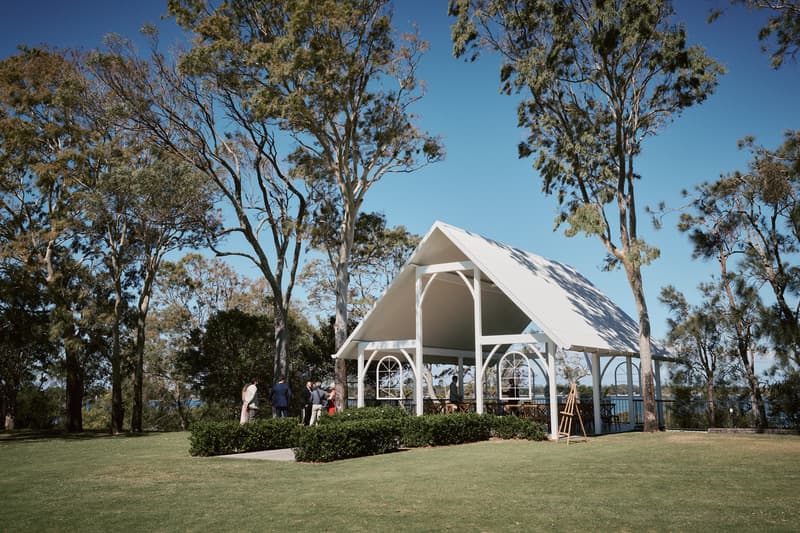 A small group of wedding guests stand near the white pavilion at Sandstone Point Hotel — The Pavilion, surrounded by trees and green lawn under a clear blue sky.