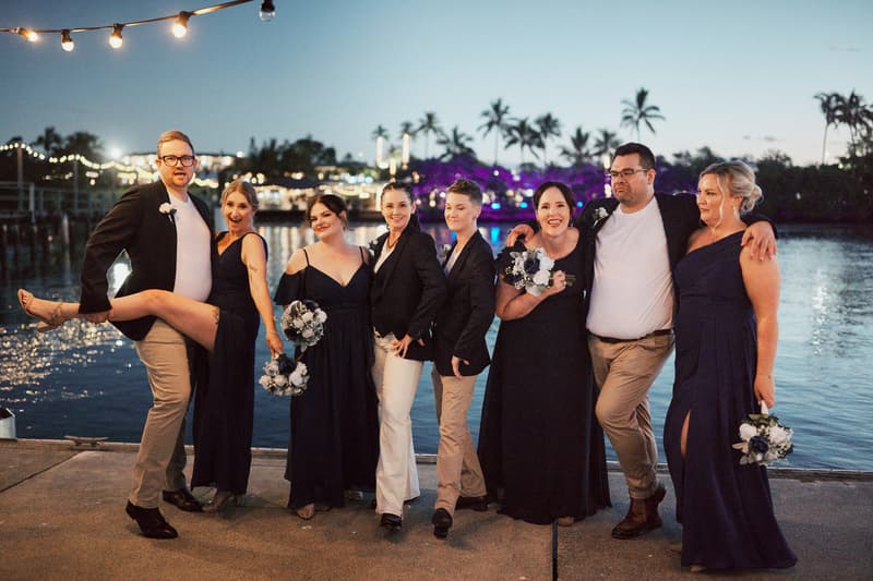 Wedding party members pose together outdoors at dusk near water with string lights overhead at Sandstone Point Hotel.