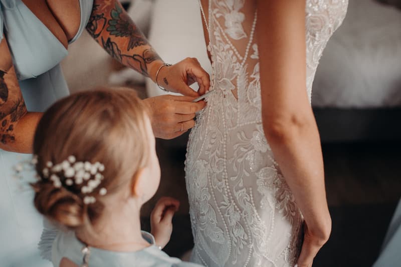 The bride is having the back of her lace wedding dress buttoned by a bridesmaid while a flower girl looks on at Sandstone Point Hotel — Pavilion.