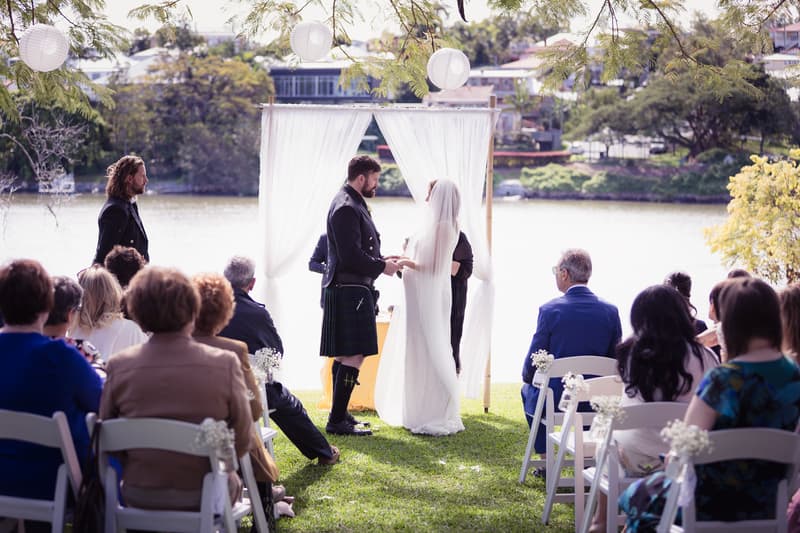 The bride Francesca and groom Ben stand facing each other holding hands under a white fabric arch at the Toowong Rowing Club by the Brisbane River, with an officiant behind them and guests seated on white chairs watching the ceremony.