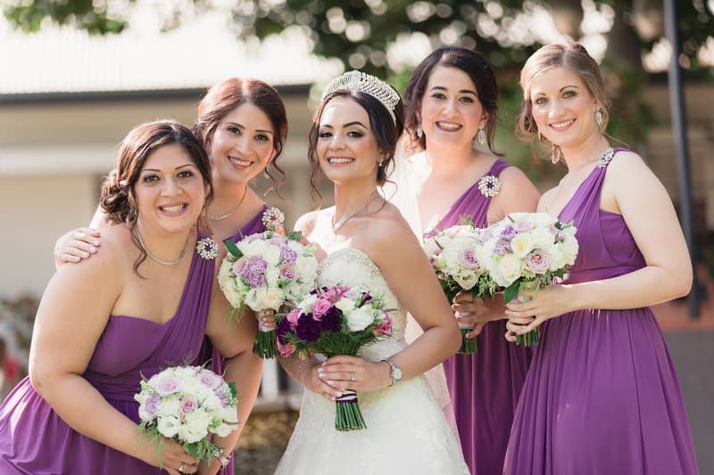 The bride Maryam stands with four bridesmaids in purple dresses, all holding bouquets of white and purple flowers, posing outdoors.