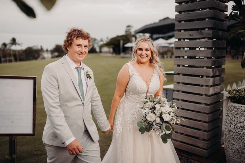 Bride Chloe and groom Brodie hold hands and smile while walking outdoors at Sandstone Point Hotel during their couple portraits session.
