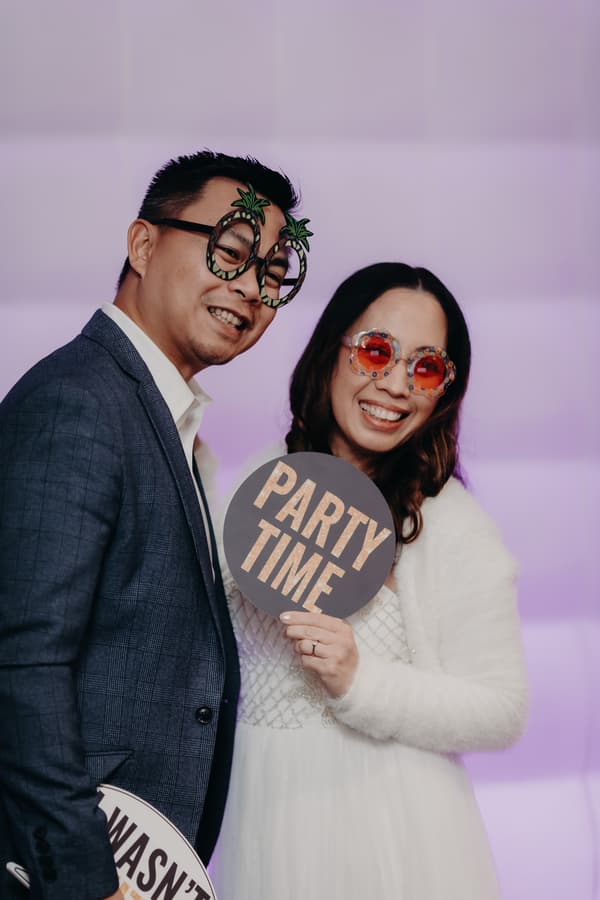 The groom Timothy and bride Henny pose together at the reception stage of Royal on the Park, wearing novelty glasses and holding signs that say 'PARTY TIME' and partially visible text.