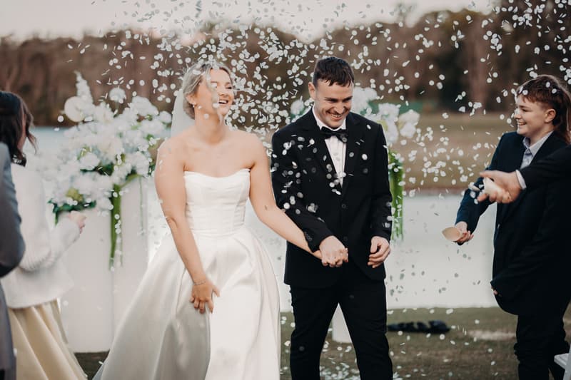 Bride Libby and groom Kyle hold hands and smile as guests throw confetti at their wedding ceremony at The Tides — The Water's Edge.
