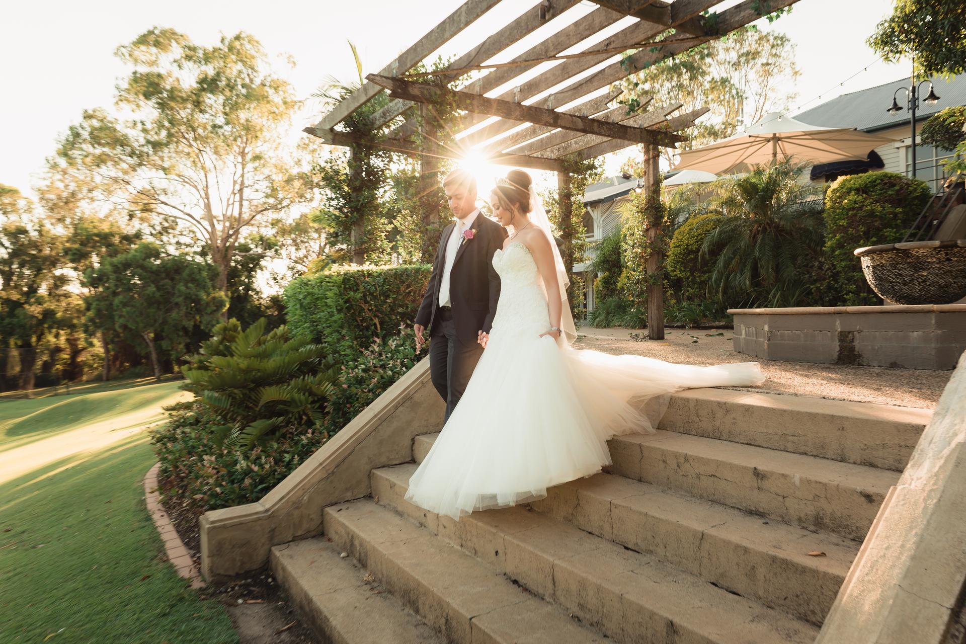 The bride Maryam and groom Pasha walk down outdoor stone steps under a wooden pergola at Hillstone St Lucia — The Quartyard with sunlight filtering through the structure.