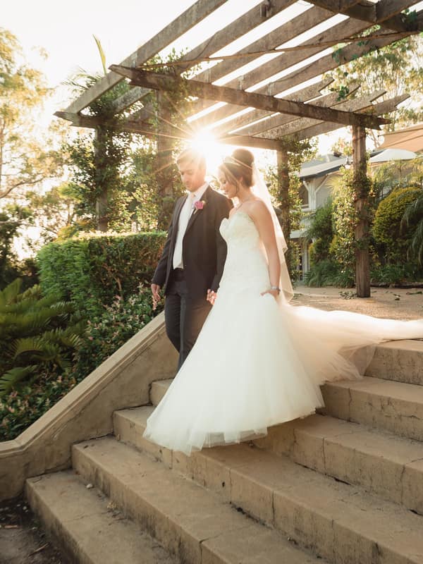 The bride Maryam and groom Pasha walk down outdoor stone steps under a wooden pergola at Hillstone St Lucia — The Quartyard with sunlight filtering through the structure.