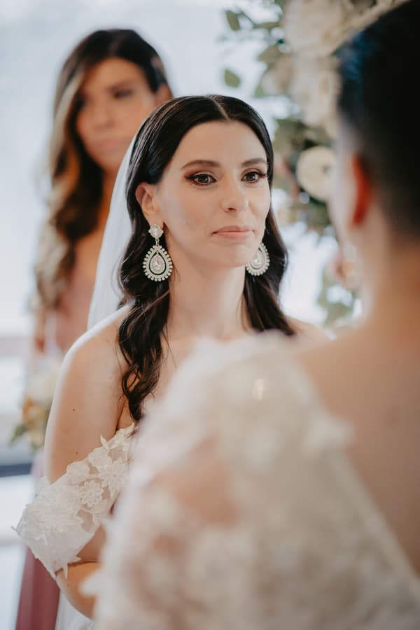 Bride with long dark hair and large earrings faces another person wearing a lace dress, with a bridesmaid blurred in the background.