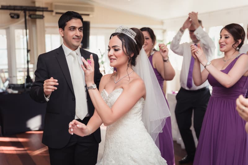 The bride Maryam in a white wedding gown and tiara dances with the groom Pasha in a black suit and white tie at Hillstone St Lucia — The Rosewood Room, surrounded by guests and bridesmaids in purple dresses also dancing.