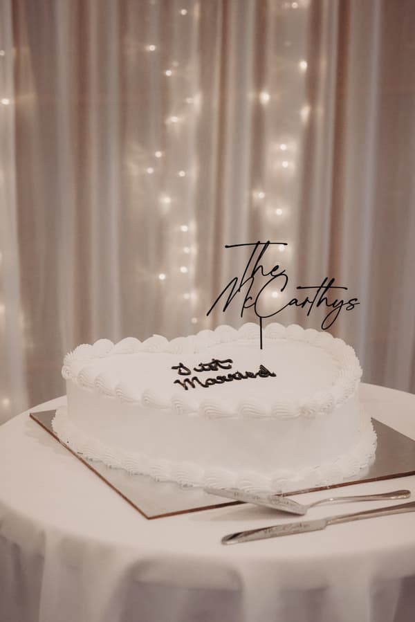 Heart-shaped white wedding cake with piped icing and black script topper reading 'The McCarthys' and 'Just Married' on a round table at The Tides — The Pandanus Room.