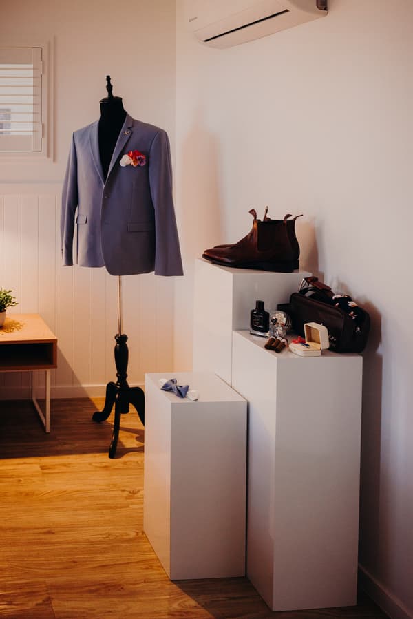 Groom's light blue suit jacket displayed on a mannequin with a boutonniere, brown leather shoes, bow tie, cologne, and other accessories arranged on white pedestals at Sandstone Point Hotel.