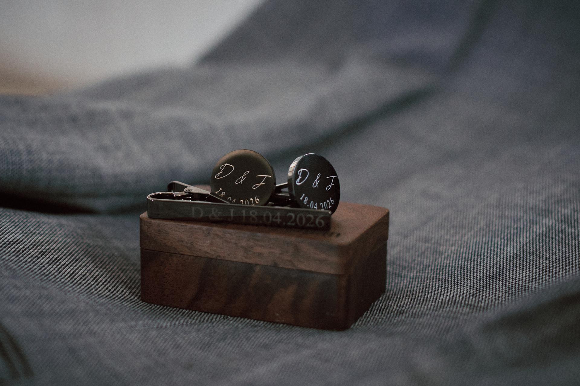 Close-up of personalized cufflinks and a tie clip engraved with 'D & J 18.04.2026' resting on a wooden box placed on fabric.