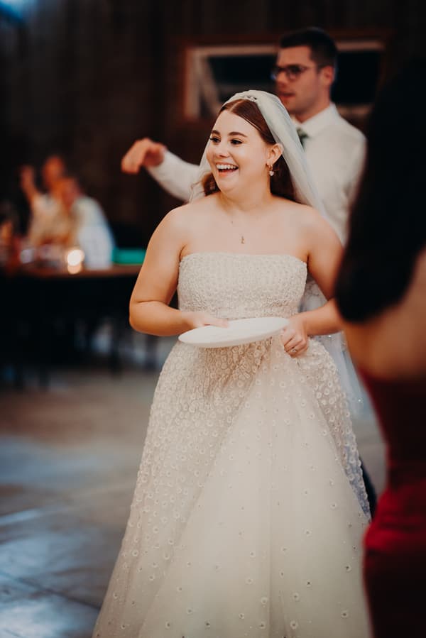 The bride Lilly holds a plate and smiles during the reception at Yabbaloumba Retreat — The Shed, with the groom Connor visible behind her.