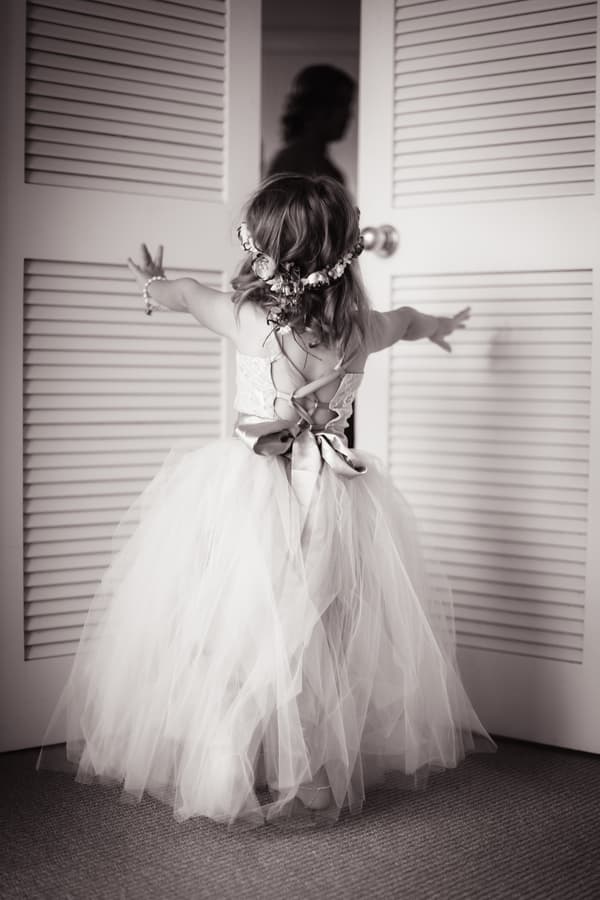 A flower girl in a tulle dress with a floral headband stands facing closed louvered doors, reaching out with both arms. A blurred silhouette of a woman is visible through the gap between the doors.
