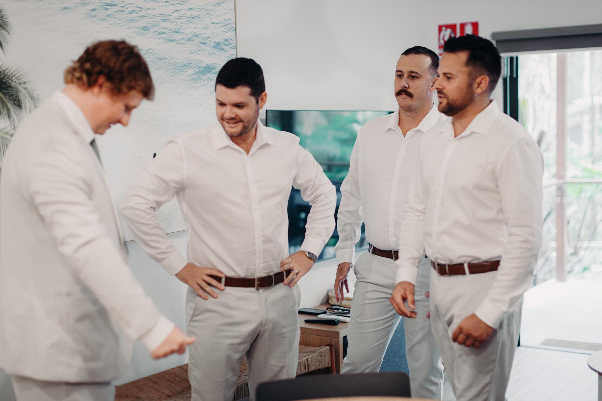 Four groomsmen dressed in white shirts and light-colored pants stand indoors at Sandstone Point Hotel, preparing or conversing before the event.