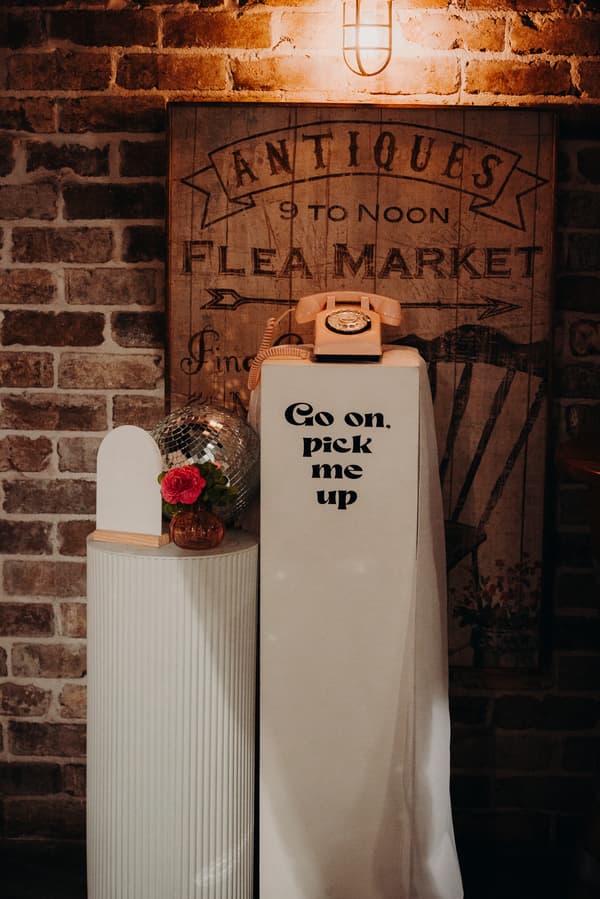 A vintage rotary telephone sits atop a white pedestal draped with a cloth that reads 'Go on, pick me up' at Sandstone Point Hotel — Cellar. Next to it is a smaller white pedestal with a small vase holding a pink flower and a disco ball, set against a brick wall with a sign reading 'ANTIQUES 9 TO NOON FLEA MARKET'.