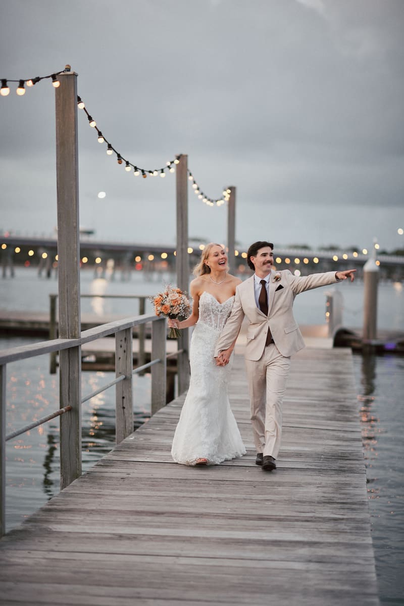 Morgan and Joel walk hand in hand on a wooden pier decorated with string lights, with water and a bridge visible in the background. What is an engagement party by She Said Yes.
