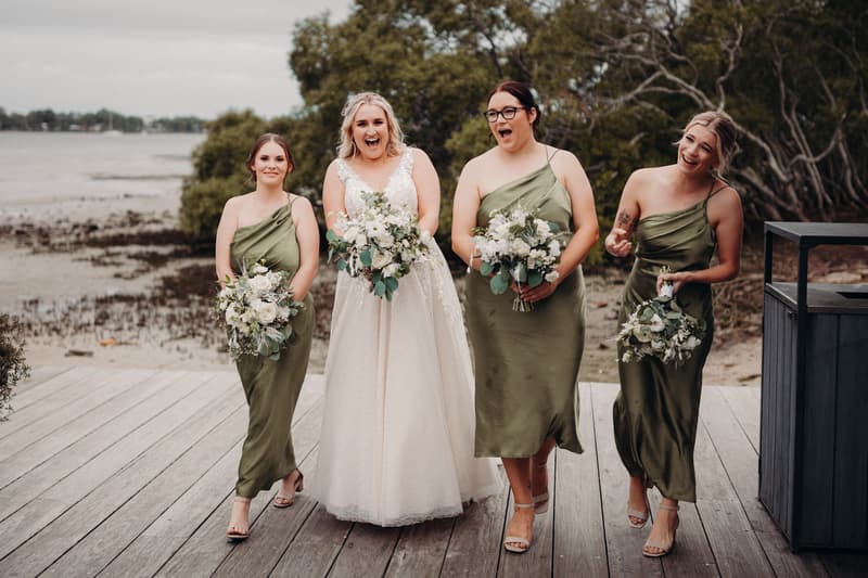 The bride Chloe in a white wedding gown walks alongside three bridesmaids in olive green dresses holding bouquets on a wooden deck at Sandstone Point Hotel with a waterfront and trees in the background.