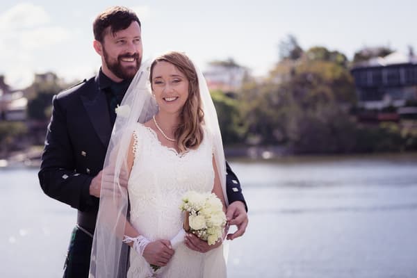 Francesca the bride and Ben the groom pose together outdoors near water at Toowong Rowing Club — The Malouf Room, with Francesca holding a bouquet of white flowers and wearing a lace wedding dress and veil, and Ben dressed in a dark suit with a tartan kilt.