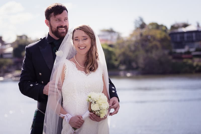 Francesca the bride and Ben the groom pose together outdoors near water at Toowong Rowing Club — The Malouf Room, with Francesca holding a bouquet of white flowers and wearing a lace wedding dress and veil, and Ben dressed in a dark suit with a tartan kilt.