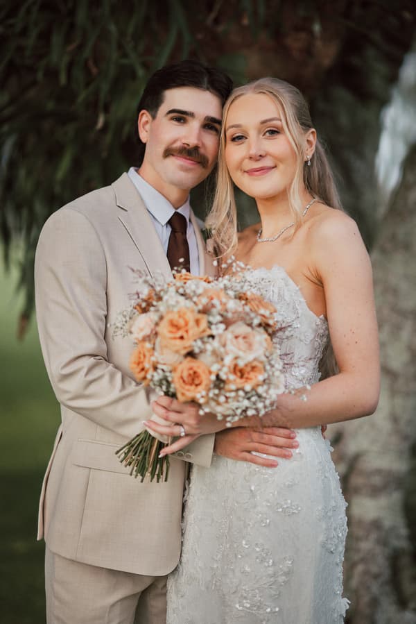 Morgan the bride and Joel the groom pose together holding a bouquet of peach and white flowers at Sandstone Point Hotel, standing in front of a tree.