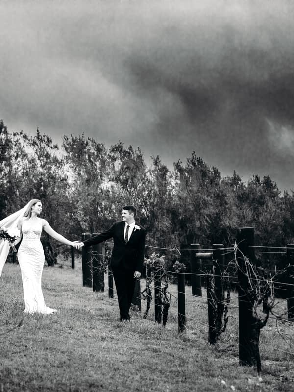 Chelsea and Brent walk hand in hand through a vineyard at Preston Peak Winery Functions Centre, with Chelsea in her wedding dress and veil and Brent in a suit.