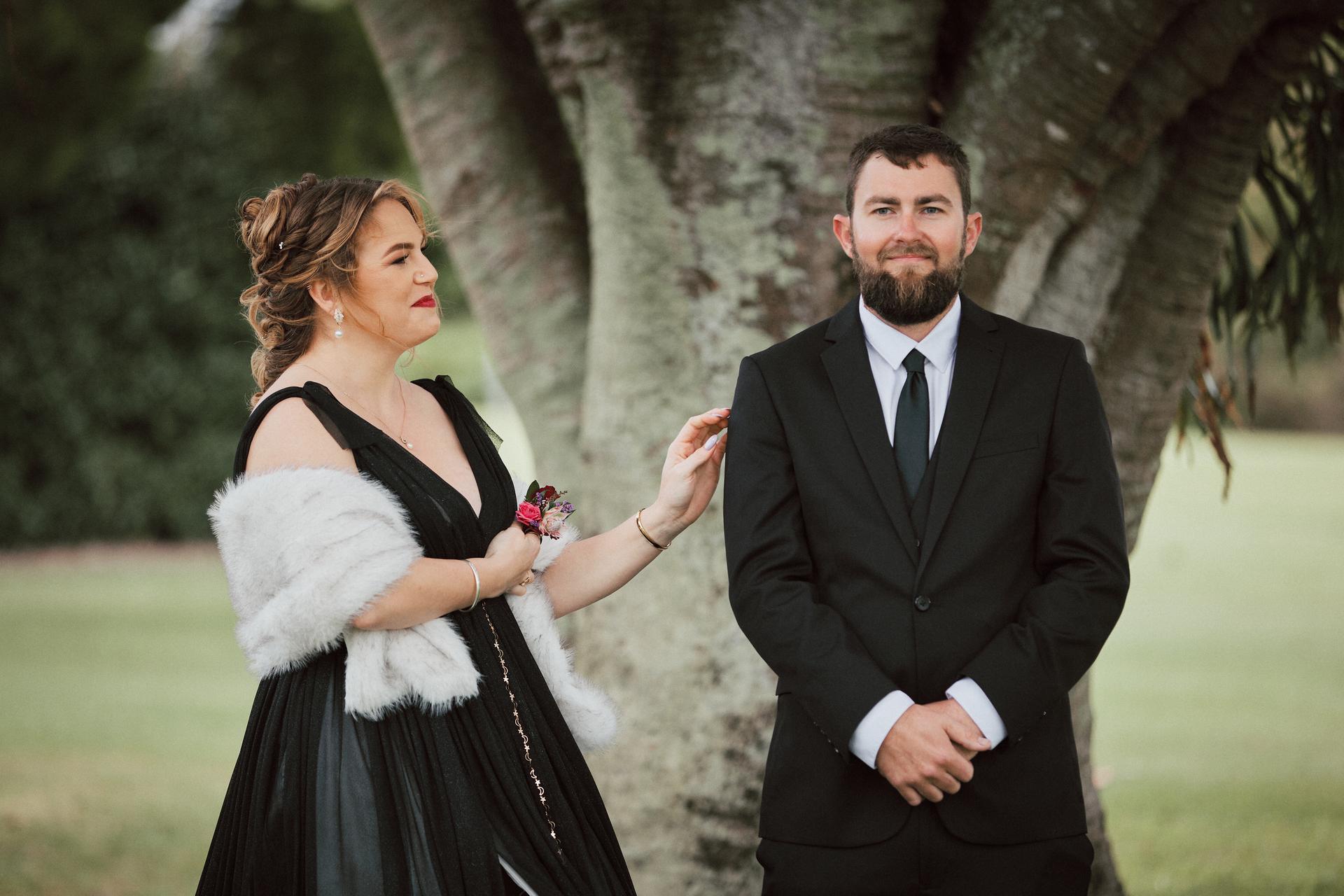 Jayme, the bride, in a black dress with a white fur stole, stands next to Christopher, the groom, who is wearing a black suit and tie, in front of a large tree at Sandstone Point Hotel — The Pavilion.