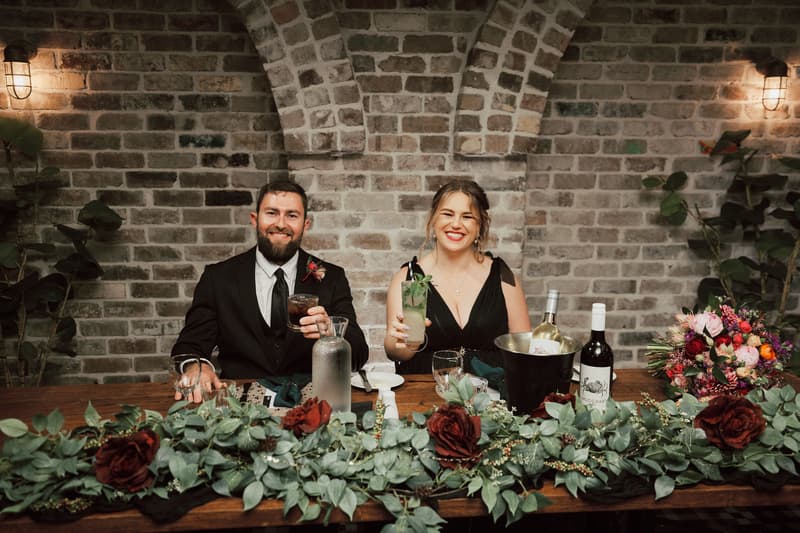 Bride Jayme and groom Christopher sit at the reception head table at Sandstone Point Hotel — The Cellar, each holding a drink. The table is decorated with greenery and red roses, with wine bottles and a floral bouquet placed on it. The background features a brick wall with two wall lights and some plants.