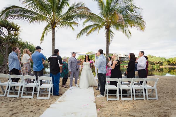 Bride Kelly and groom Shane stand together on a white aisle runner at Novotel Sunshine Coast Resort beach ceremony site, surrounded by guests standing and seated on white chairs under palm trees near a water body.