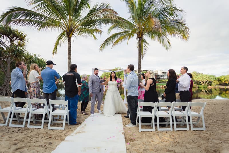 Bride Kelly and groom Shane stand together on a white aisle runner at Novotel Sunshine Coast Resort beach ceremony site, surrounded by guests standing and seated on white chairs under palm trees near a water body.