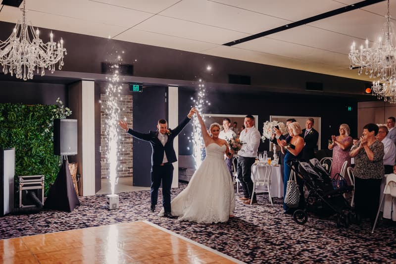Bride Emily and groom Dylan enter the reception stage at Sandstone Point Hotel — Pumicestone Room, holding hands with sparklers ignited behind them as guests applaud.