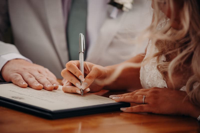 The bride Chloe signs a document at the Sandstone Point Hotel — Pavilion, with the groom Brodie's hands resting on the table nearby.