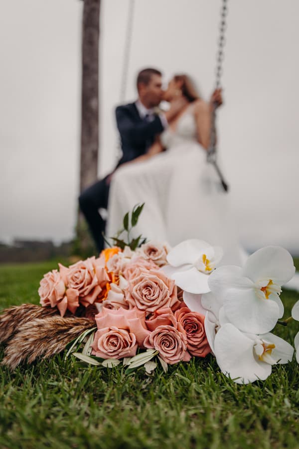 Chelsea and Brent sit on a swing at Preston Peak Winery Functions Centre with a bouquet of pink roses and white orchids in the foreground on the grass.