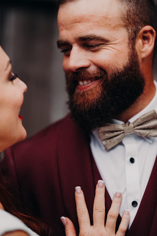 The bride and groom share a close moment, with the bride's hand resting on the groom's chest. The groom wears a burgundy suit jacket, white shirt, and patterned bow tie.