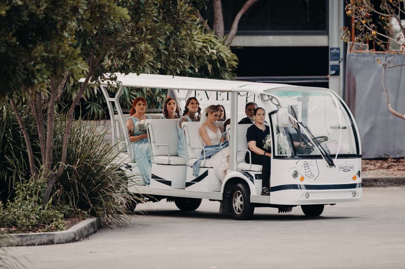 The bride Courtney and bridesmaids are seated in a white open-sided shuttle vehicle at Sandstone Point Hotel — Pavilion, accompanied by two other adults in the front seats.