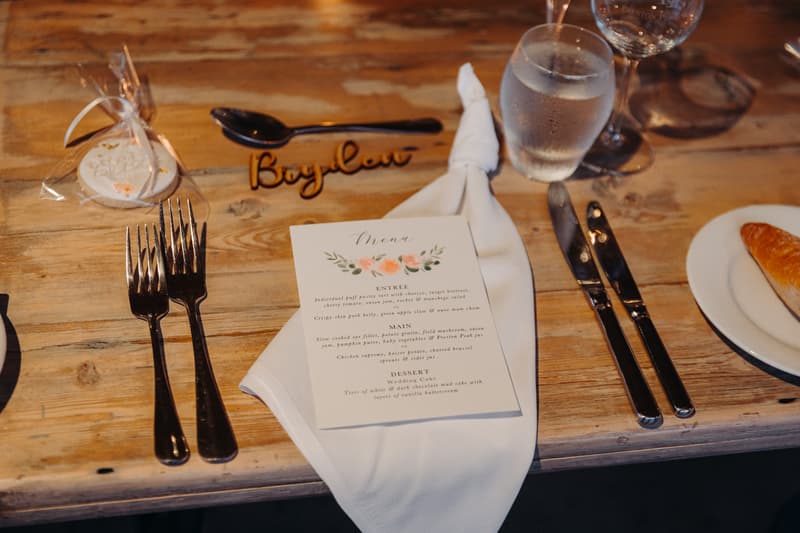 Wedding reception table setting at Preston Peak Winery Functions Centre with a menu card, cutlery, glass of water, wine glass, a croissant on a plate, and a name tag reading 'Baylou'.