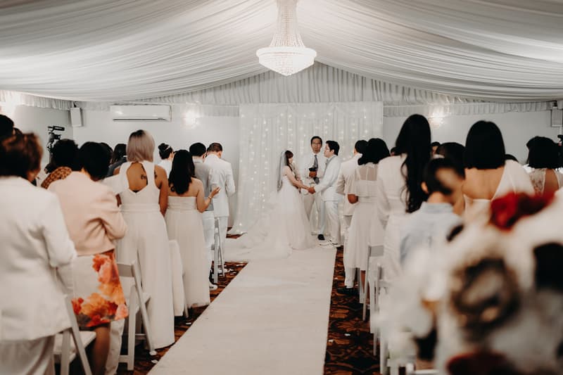 Bride Henny and groom Timothy hold hands facing the officiant during their wedding ceremony at Royal on the Park, with guests seated on either side of the aisle.