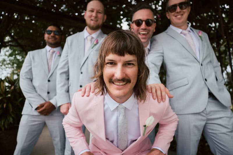 Jake, the groom, in a pink suit with a white rose boutonniere, poses in front of four groomsmen wearing light grey suits with pink ties and boutonnieres at Eatons Hill Hotel — Lakeside.