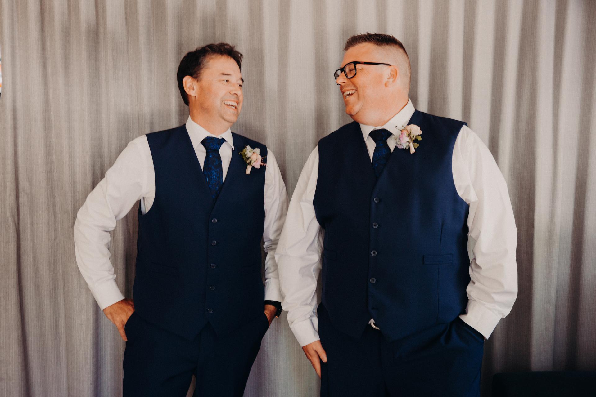 Two men in matching navy vests, white shirts, and ties stand indoors in front of a gray curtain, smiling and looking at each other.