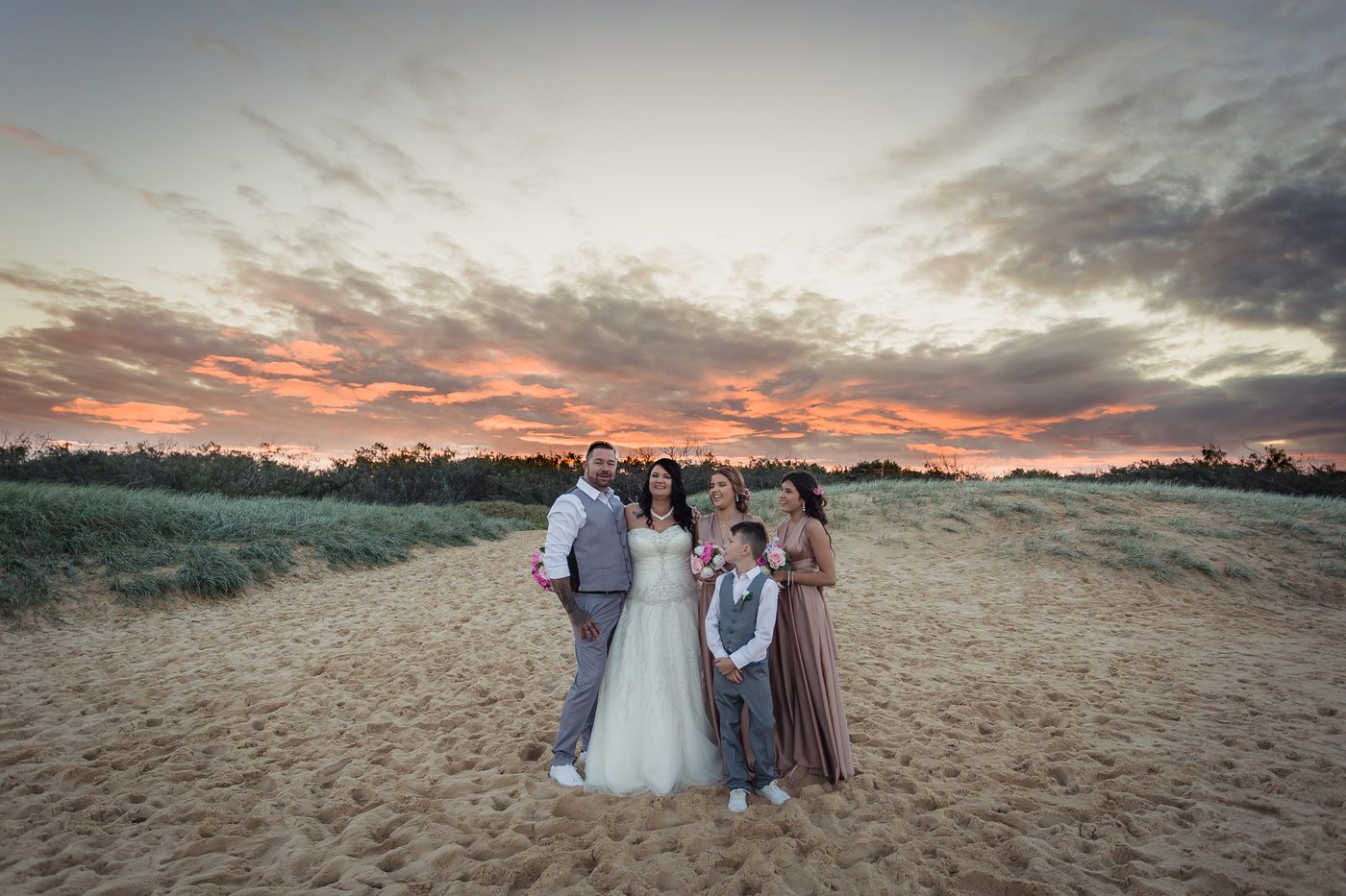 Kelly the bride, Shane the groom, two bridesmaids, and a young boy in a suit pose together on a sandy beach at sunset near. Wedding photography contracts by She Said Yes.