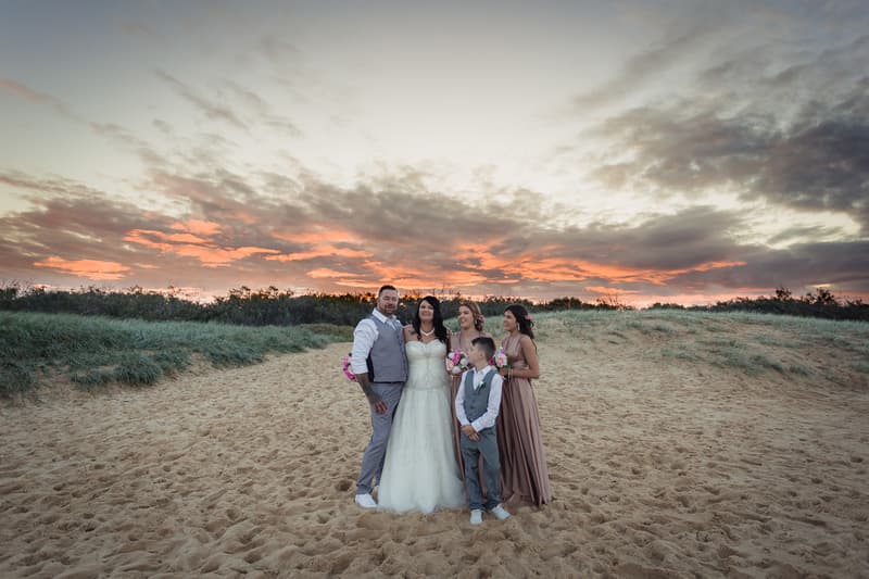 Kelly the bride, Shane the groom, two bridesmaids, and a young boy in a suit pose together on a sandy beach at sunset near Novotel Sunshine Coast Resort.
