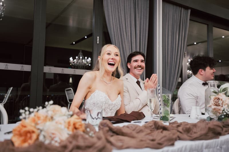 The bride Morgan and groom Joel sit at the head table in The Pumicestone Room at Sandstone Point Hotel during the wedding reception, smiling and clapping. Another male guest in a white shirt and bow tie is seated beside them.