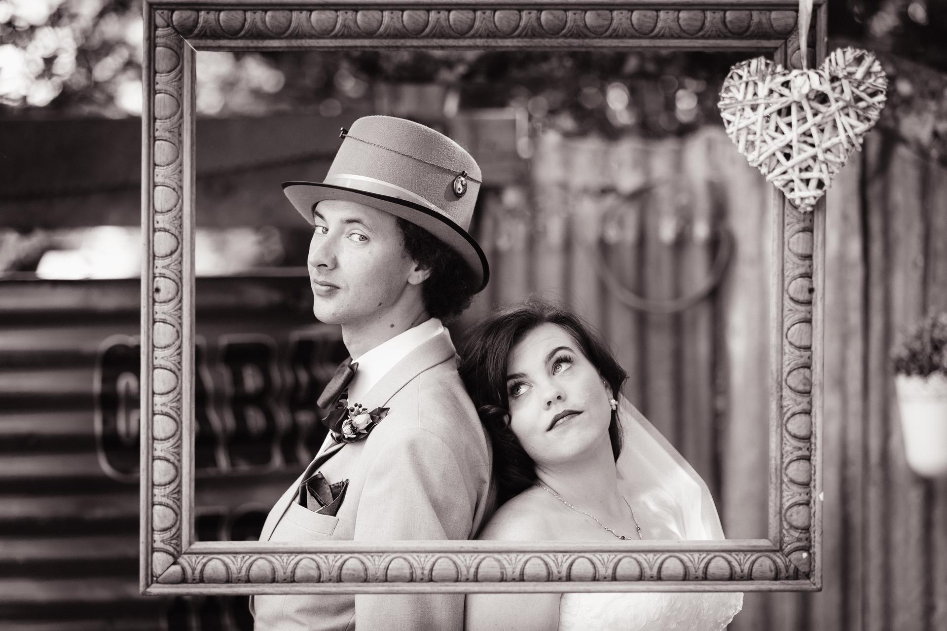 The bride Holly and groom Paul pose back-to-back inside a large decorative frame at Kwila Lodge, with a heart-shaped wicker decoration hanging on the frame's top right corner.