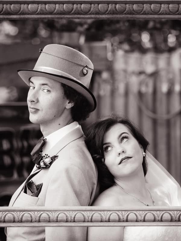 The bride Holly and groom Paul pose back-to-back inside a large decorative frame at Kwila Lodge, with a heart-shaped wicker decoration hanging on the frame's top right corner.
