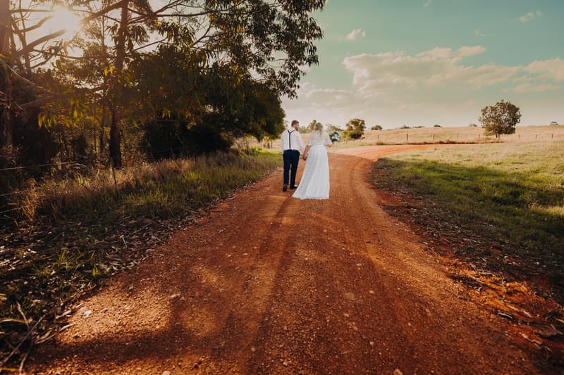 The bride Annie and groom Trent walk hand in hand down a dirt road at Ocean View Estates, surrounded by trees and open fields under a partly cloudy sky.