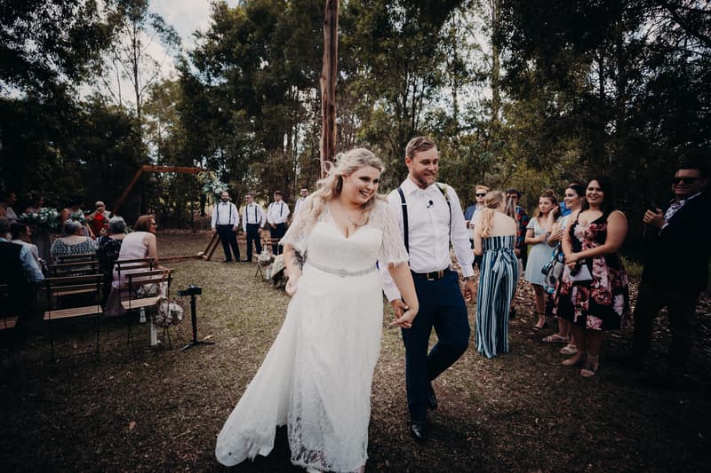 Bride Annie and groom Trent walk hand in hand down the aisle at Ocean View Estates after their wedding ceremony, with guests seated and standing on either side and groomsmen in suspenders standing near the altar.